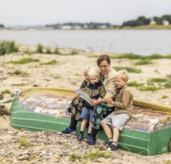 a woman sits on an upside-down boat on a beach with three little boys